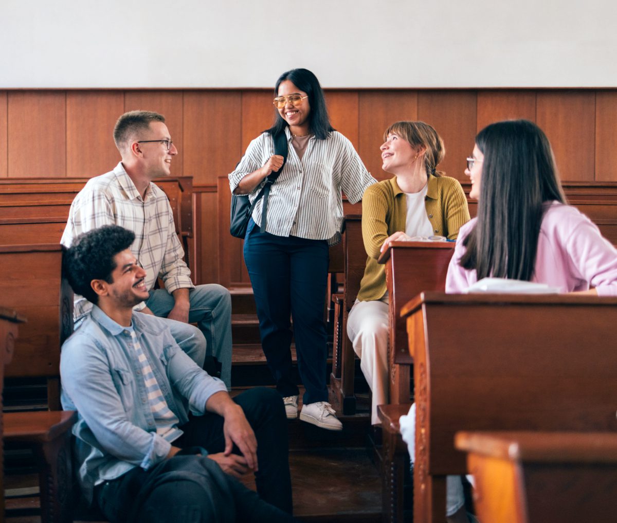A group of university students gather in a lecture hall, chatting and smiling. Their diverse backgrounds highlight inclusiveness and camaraderie, portraying a positive learning environment and mutual support.
