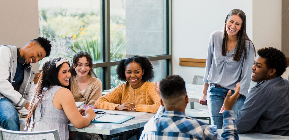 The multi-ethnic group of classmates and their mid adult female teacher smile and laugh when the unrecognizable teenage boy makes a joke.