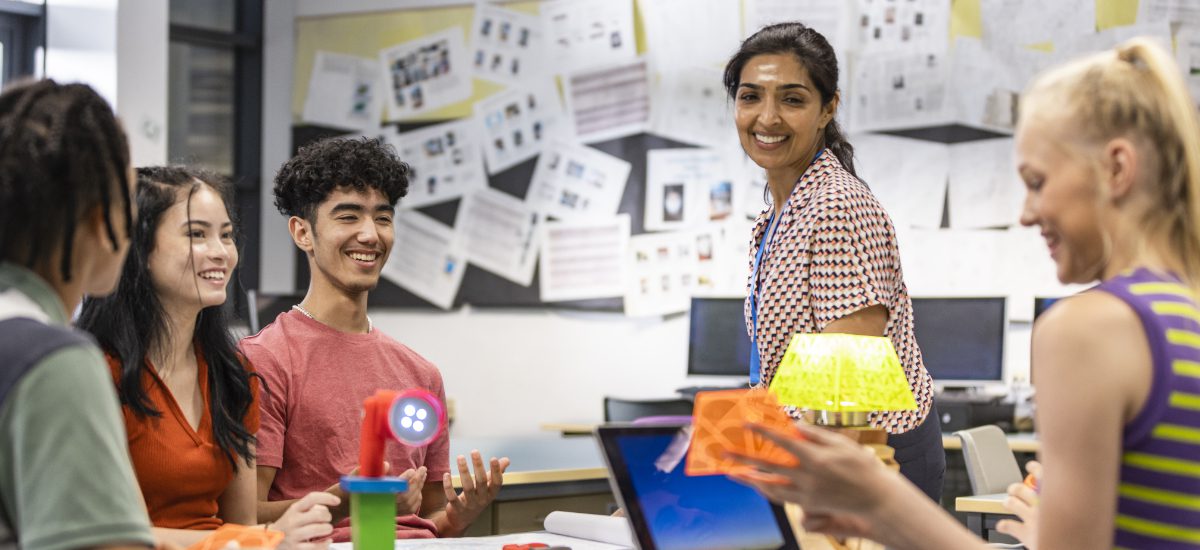 A shot of a group of students with their teacher in class, discussing their work. They are all wearing casual clothing and smiling, sitting in a classroom.